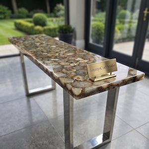 May include: A rectangular console table featuring a polished surface composed of numerous brown, tan, and cream-colored agate slices embedded in resin. The table is supported by two silver-toned metal U-shaped legs and displays a gold-toned nameplate reading JimmysCraftStudio.