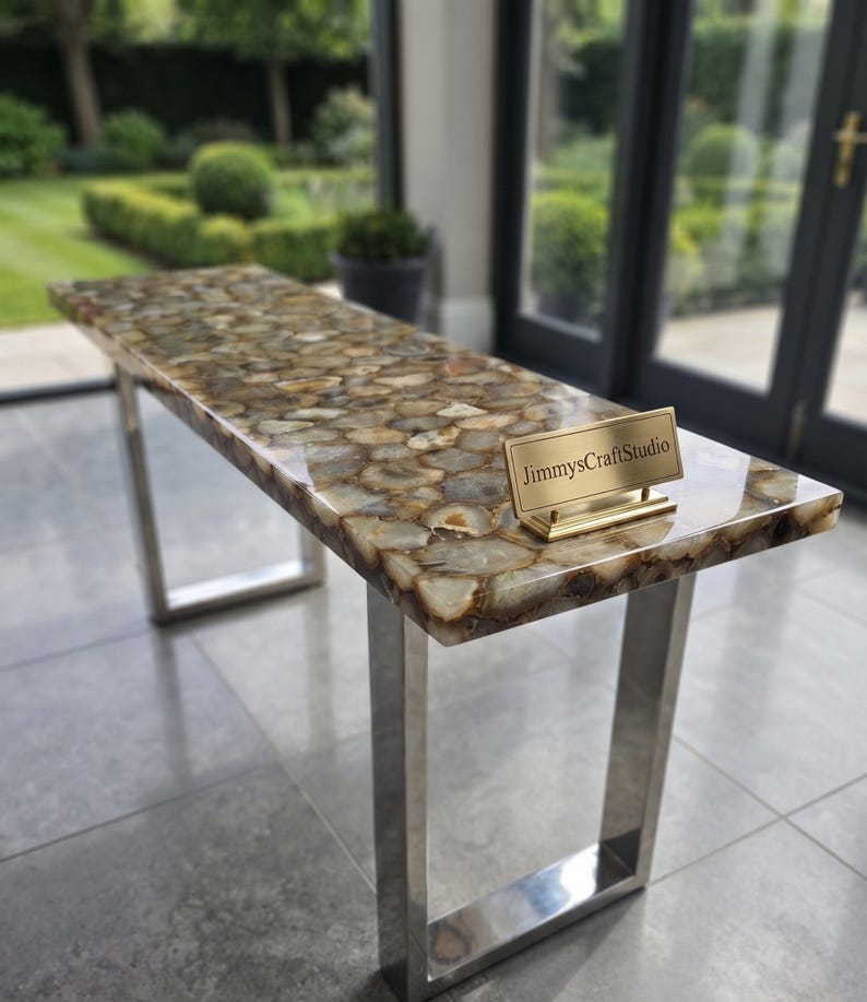 May include: A rectangular console table featuring a polished surface composed of numerous brown, tan, and cream-colored agate slices embedded in resin. The table is supported by two silver-toned metal U-shaped legs and displays a gold-toned nameplate reading JimmysCraftStudio.
