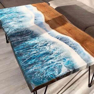 May include: A rectangular coffee table with a unique design. The table features a resin top with a blue and white ocean wave pattern, transitioning to a natural wood section. The table has black metal hairpin legs, creating a modern aesthetic.
