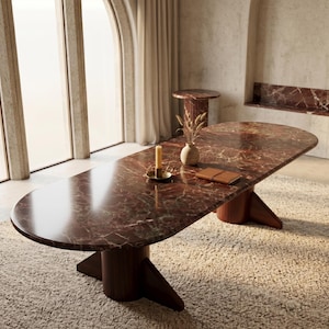 Op de afbeelding: Een ovale marmeren tafel in roodbruin met een donkere houten voet. De tafel staat in een kamer met neutrale tinten. Een kaars, vaas en notitieboek liggen op de tafel. De tafel is ongeveer 244 cm lang. De kamer heeft grote ramen en een tapijt.