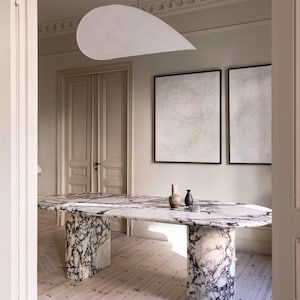Pode incluir: Uma mesa de jantar em mármore com um padrão de veios em preto e branco. A mesa tem um tampo retangular arredondado e duas bases de pedestal cilíndricas. Dois pequenos vasos estão sobre a mesa. A sala tem paredes claras e chão de madeira.