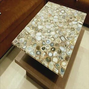 May include: A rectangular coffee table with a mosaic top made of polished, irregularly shaped stones in shades of beige, gray, and brown. The table has a dark brown wooden base. The table is a decorative furniture piece.