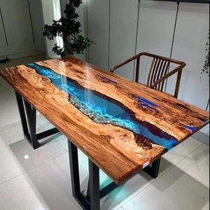Peut inclure: Une table rectangulaire en bois avec une rivière de résine bleue traversant le centre. La table a un fini grain de bois naturel avec des pieds en métal noir. La rivière de résine est remplie de petites pierres décoratives.