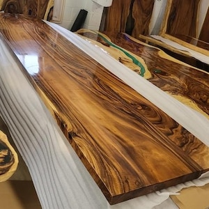 May include: A rectangular wooden table top with a polished surface. The wood grain displays a deep brown hue with lighter brown and tan streaks. The table top is resting on a white protective covering. Other similar pieces are visible in the background.