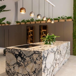 Puede incluir: Isla de cocina moderna con encimera de mármol con vetas blancas y negras. Encima, una hilera de lámparas colgantes con pantallas de cristal. La cocina también incluye gabinetes marrones oscuros, vitrinas y plantas en macetas.