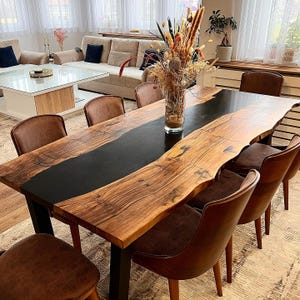 May include: A large, handcrafted dining table with a live edge wooden surface and a black epoxy resin center. The table is surrounded by brown leather chairs. A vase of dried flowers sits in the center of the table.