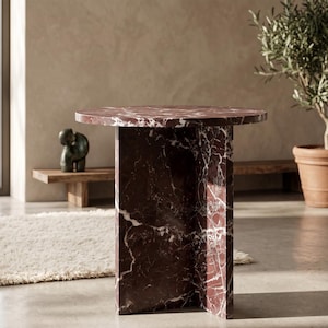 May include: A modern, red marble side table with a round top and a rectangular base. The table has white veining throughout the red marble. The table is a stylish piece of furniture for a living room or bedroom.
