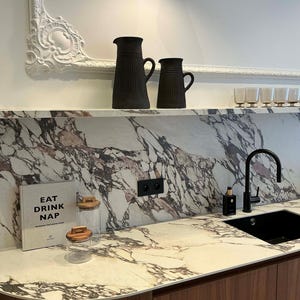 Puede incluir: Escena de cocina con encimera de mármol y fregadero negro. Dos jarras negras sobre una estantería. Un libro con el texto "EAT DRINK NAP" es visible, junto con tarros de cristal y un frasco de perfume.