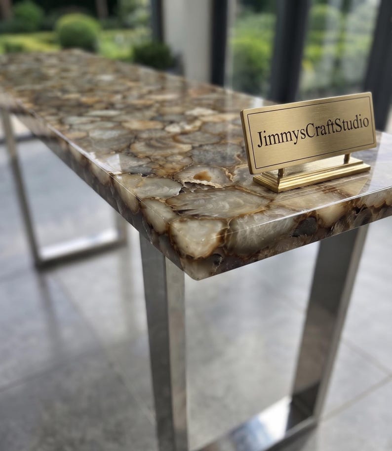 May include: A rectangular console table featuring a polished surface made of multi-tonal brown and cream agate slices. The table has a metallic silver frame and a small gold-toned rectangular plaque with the text JimmysCraftStudio.