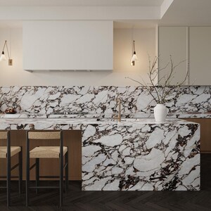 Op de afbeelding: Moderne keuken met een marmeren aanrechtblad en achterwand met donkere adering. Het eiland heeft een houten basis en wordt gecombineerd met barkrukken met geweven zittingen. Boven hangen hanglampen en een witte afzuigkap. Een vaas met takken staat op het aanrecht.