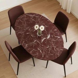 Può includere: Vista dall'alto di un set da pranzo con un tavolo ovale bordeaux con motivo marmorizzato, circondato da quattro sedie abbinate. Un piccolo vaso con fiori bianchi è sul tavolo. Le sedie hanno gambe nere e sedute in tessuto bordeaux.