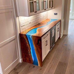 May include: A custom-made wooden countertop with a vibrant blue resin river design. The countertop is installed on white cabinets with black hardware. The wood has a natural, warm tone, contrasting with the cool blue resin. The countertop is part of a kitchen or bar area.