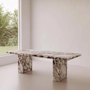 Puede incluir: Una mesa rectangular de mármol con un patrón blanco y burdeos. La mesa tiene una tapa gruesa y dos patas rectangulares. La mesa está en una habitación con una ventana grande y paredes blancas.