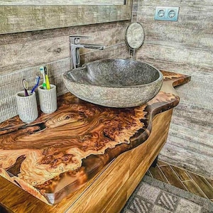 Puede incluir: Un mueble de baño con encimera de madera natural y un lavabo de piedra gris. La encimera presenta un patrón de vetas de madera único con un acabado de resina transparente. Se ven dos cepilleros y un grifo cromado.
