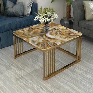 May include: A square coffee table featuring a polished agate stone tabletop with brown, gold, and white marbled patterns. The table is supported by a gold-toned metal frame constructed with vertical bars on each side, creating an open, airy structure.