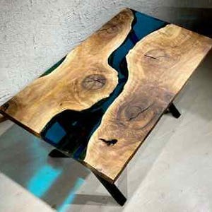 May include: A rectangular coffee table with a live edge design. The table features a natural wood surface with a river of blue resin. The table is supported by black metal legs. The table measures approximately 48 inches long and 24 inches wide.