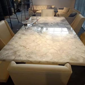 May include: A rectangular dining table featuring a polished, translucent white and cream-colored stone slab top with a natural, irregular mosaic pattern. The table is surrounded by several upholstered, light-colored chairs with high backrests.