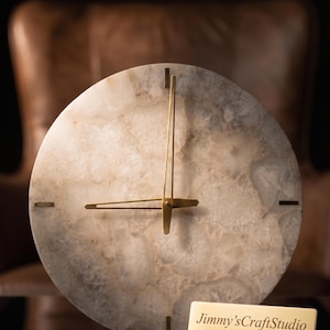 May include: A round wall clock with a light beige and gray marbled face. The clock has gold-colored hands and markers at each hour. The clock face is made of a natural stone material. The clock is displayed in front of a blurred brown background. The clock has the text "Jimmy's CraftStudio".