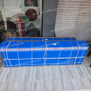 May include: A large, rectangular package wrapped in blue paper and secured with white straps. The package is labeled with "FRAGILE" tape. The background shows a storefront with various decorative items.