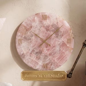 Puede incluir: Un reloj de pared circular hecho de piedra de cuarzo rosa translúcido, con números romanos dorados en las posiciones de las doce, tres, seis y nueve, manecillas doradas delgadas y una placa dorada con la etiqueta Jimmy'sCraftStudio.