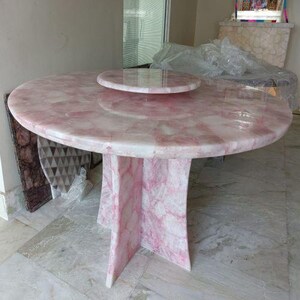 May include: A round dining table with a rose quartz top and a smaller, matching lazy susan. The table has a pedestal base with four legs. The pink and white stone has a marbled appearance.