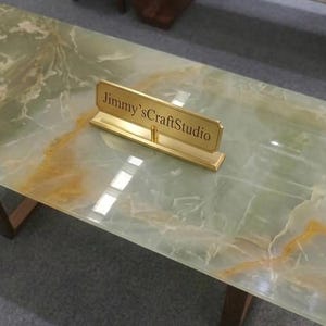 Op de afbeelding: Een rechthoekige salontafel met een gepolijst lichtgroen en amberkleurig geaderd stenen blad. De tafel heeft een goudkleurige metalen naamplaatstandaard in het midden met de tekst Jimmy'sCraftStudio erin gegraveerd, op donkerhouten poten.