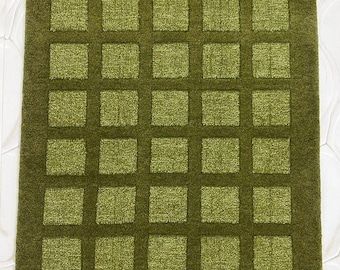 May include: A square, olive green rug with a grid pattern of darker green squares. The rug has a textured appearance, with a darker border surrounding the grid design. The rug is placed on a white, patterned surface.