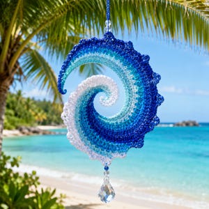 May include: A spiral-shaped crochet ornament featuring a gradient of blue and white yarn, mimicking the appearance of a crashing ocean wave. The piece is suspended by a blue string and features a clear, faceted crystal drop at the bottom.