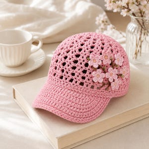 May include: A light pink crochet baseball cap featuring an open-weave pattern on the crown and a solid brim. The side of the cap is decorated with several small, layered pink and white crocheted flower appliqués with yellow centers.