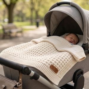 Peut inclure: Une couverture pour bébé de couleur crème en tricot gaufré avec une bordure côtelée et une étiquette en similicuir marron portant l'inscription "HAND KNITTED WITH LOVE". La couverture recouvre un bébé dans une poussette grise.