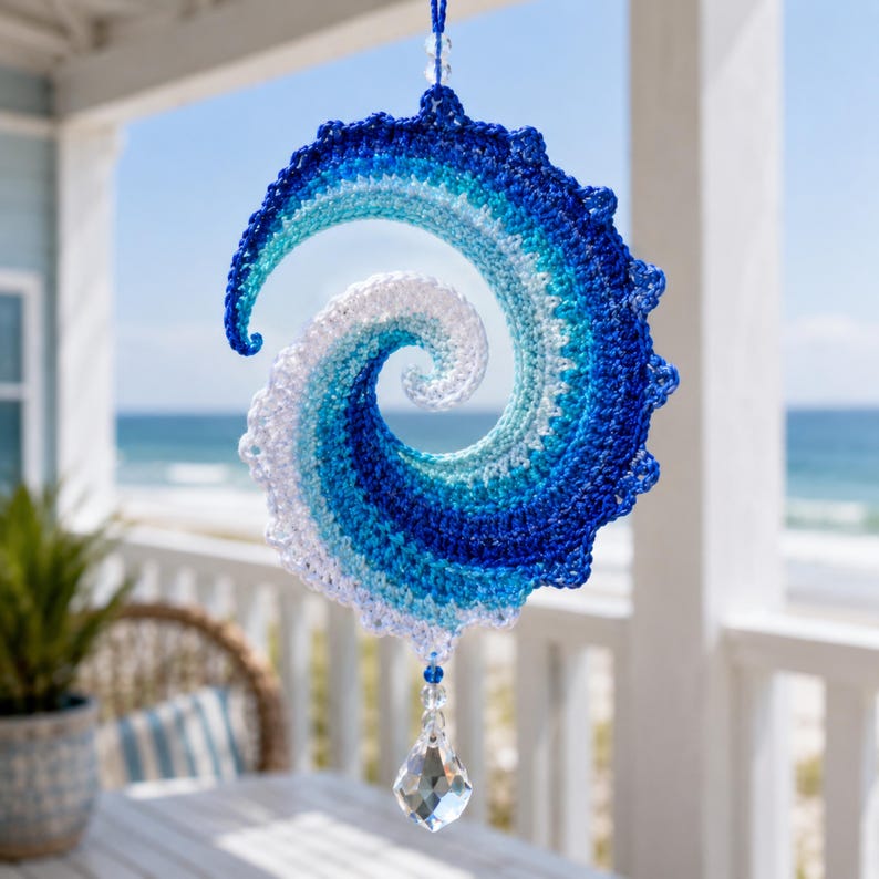 May include: A spiral-shaped crochet hanging ornament featuring a gradient of blue and white yarn, mimicking the appearance of a crashing ocean wave. The piece is suspended by a blue string and features a clear, faceted crystal drop at the bottom.