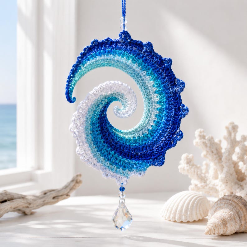 May include: A spiral-shaped crochet ornament featuring a gradient of blue, turquoise, and white yarn. The piece is suspended by a blue string and features a clear, faceted crystal drop hanging from the bottom point of the spiral structure.