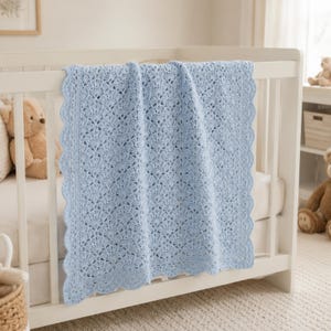 May include: A light blue crochet baby blanket featuring an intricate shell stitch pattern throughout the center and a decorative scalloped border along all four edges. The blanket is draped over the side of a white wooden crib.