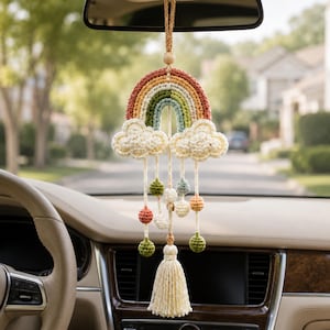 Peut inclure: Une décoration de voiture en crochet représentant un arc-en-ciel avec des bandes terracotta, moutarde et vert sauge, encadré par deux nuages crème. Des fils crème pendent avec des petites sphères crochetées et un grand pompon crème.