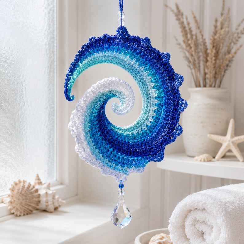 May include: A spiral-shaped crochet hanging ornament featuring a gradient of blue, teal, and white yarn. The piece is suspended by a blue string and features a clear, faceted crystal drop at the bottom, with a small clear bead at the top.