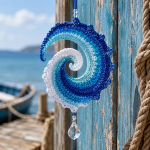 May include: A spiral-shaped crochet ornament featuring a gradient of blue and white yarn, mimicking the appearance of a crashing ocean wave. The piece is suspended by a blue string and features a clear, faceted crystal drop at the bottom.