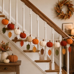 Op de afbeelding: Een decoratieve slinger met afwisselend gehaakte pompoenen in tinten oranje, rood en crème, afgewisseld met gehaakte herfstbladeren in groen, geel en bruin, allemaal geregen aan een lichtgekleurd gedraaid koord.