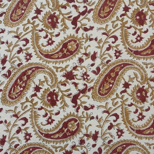 May include: Close-up of a fabric with a repeating paisley pattern. The design features large, teardrop-shaped paisley motifs in a deep red color, outlined in gold. The background is a cream color, with smaller floral and vine elements.