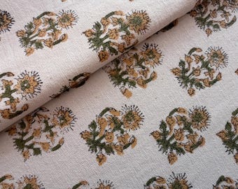 May include: Cream-colored fabric with a repeating floral pattern. The flowers are a mix of gold and green, with detailed petals and leaves. The fabric is folded, showing the texture and design.