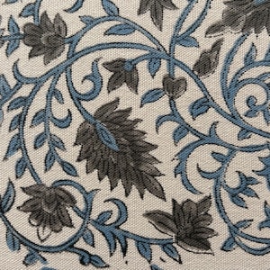 Op de afbeelding: Close-up van een stof met een herhalend bloemenpatroon. Het ontwerp toont gestileerde grijze bloemen en bladeren verbonden door blauwe wijnranken en krullen, tegen een crèmekleurige achtergrond.