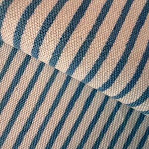 May include: Close-up of a folded fabric with a repeating pattern of blue stripes on a cream background. The fabric appears to be woven, with a textured surface. The stripes are evenly spaced and run diagonally across the image.