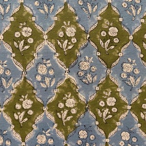 Op de afbeelding: Close-up van een stof met een herhalend bloemmotief. Het ontwerp toont witte bloemen en bladeren tegen een achtergrond van olijfgroene en lichtblauwe ruitvormen. De algehele esthetiek doet denken aan een vintage textiel.