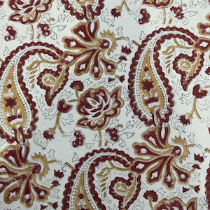 May include: Close-up of a white fabric with a repeating pattern of paisley and floral designs. The paisley elements are outlined in a dark red color and filled with a mustard yellow hue. The floral elements are dark red.
