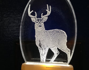 May include: A white night light with a clear oval-shaped cover featuring an etched image of a deer with antlers. The night light is illuminated with a warm white glow.