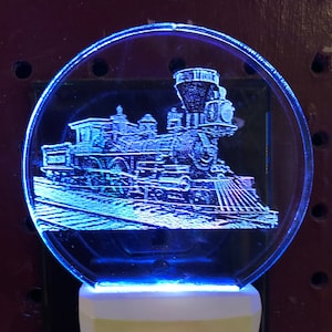 May include: A blue light illuminates a clear glass nightlight with an etched image of a vintage steam locomotive. The nightlight is plugged into an electrical outlet.