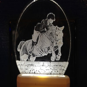 May include: A white night light with a clear plastic cover featuring a silhouette of a person riding a horse jumping over a fence. The night light is plugged into an electrical outlet.