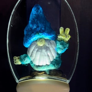 May include: A blue and white gnome figurine with a peace sign hand gesture, inside a clear glass dome nightlight. The nightlight is white and has a light bulb inside.