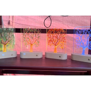 以下が含まれることがあります： 木製の土台が付いたアクリル製のランプが4つあり、それぞれ異なる色の木のデザインが施されています。木は緑、オレンジ、赤、青です。