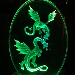 May include: A green glass oval-shaped plaque with two dragons etched into the surface. The dragons are facing each other with their tails intertwined. The plaque is illuminated from behind, making the dragons glow.