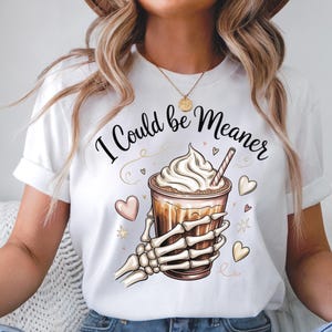 May include: White t-shirt featuring a graphic of a coffee drink with whipped cream, held by a skeleton hand. The text "I Could be Meaner" is above the coffee. The design includes hearts and stars.